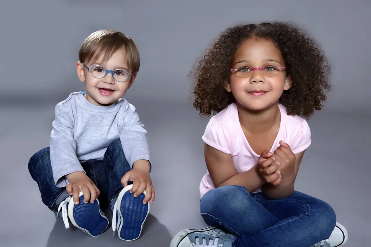 Zwei Kinder mit modischen Brillen sitzen lächelnd auf dem Boden vor grauem Hintergrund